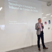 Um homem vestindo terno cinza e camisa rosa fala ao microfone em frente a uma projeção de slide.