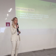 Uma mulher vestindo um terno claro fala ao microfone em frente a uma projeção de slides.