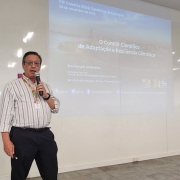 Um homem de camisa listrada fala ao microfone em frente a uma projeção de slides com o texto: O Comitê Científico de Adaptação e Resiliência Climática