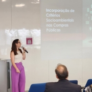 Uma mulher de camisa branca e calças cor-de-rosa fala ao microfone em frente a uma apresentação com as palavras Incorporações de Critérios Socioambientais nas Compras Públicas