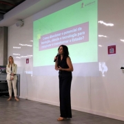 Uma mulher de vestido preto fala ao microfone em frente a uma apresentação com as palavras Como direciona o potencial da inovação, ciência e tecnologia para recuperar e transformar o Estado?