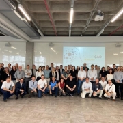 Cerca de 60 pessoas arrumadas em frente a uma apresentação em slides estão posando para a câmera, os da frente abaixam-se apoiando um joelho no chão para permitir que os de trás também sejam fotografados.