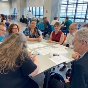 Oito pessoas conversam em torno de uma mesa quadrada. Folhas brancas com tabelas e textos estão distribuídas sobre a mesa.