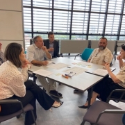 Sete pessoas conversam em torno de uma mesa quadrada. Folhas brancas com tabelas e textos estão distribuídas sobre a mesa. À direita, uma mulher gesticula enquanto fala. À esquerda, duas pessoas ouvem com a mão no queixo.