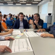 Cinco pessoas conversam em torno de uma mesa quadrada. Sobre a mesa, post-its coloridos e duas cópias em papel branco de uma tabela ou canvas organizacional.