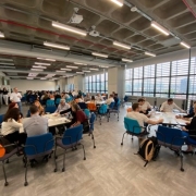 Visão de um salão amplo e de teto alto. À direita as janelas estão abertas para a luz de fora entrar. Na sala, temos uma dezena de mesas, cada uma cercada por seis ou oito pessoas sentadas conversando e discutindo sobre anotações e folhas de papel.