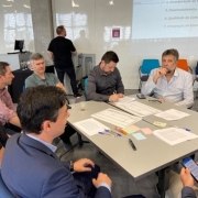 Sete pessoas conversam sentados em torno de uma mesa quadrada com folhas, tabelas, post-its coloridos e canetas espalhadas sobre a mesa.