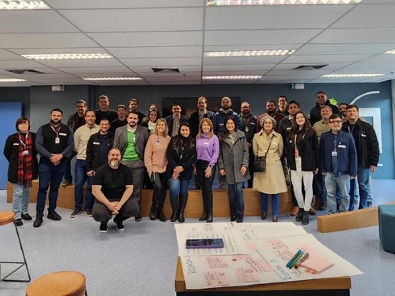 Cerca de 30 pessoas posam para fotografia. A primeira fila está de pé no chão da sala. A fila de trás está de pé sobre um palco para poderem ser fotografados.