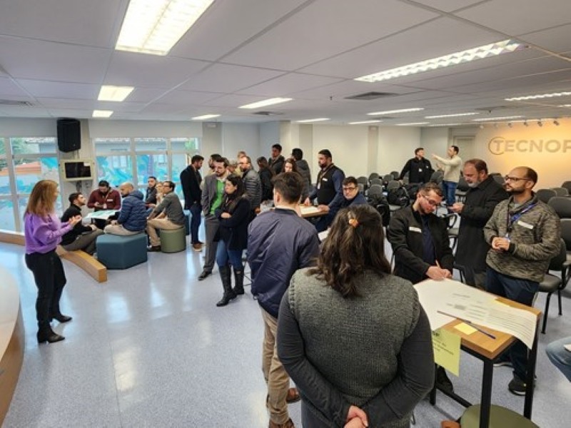 Um sala com muitas pessoas distribuídas em grupos, mesas, cadeiras e de pé conversando sobre folhas e papéis coloridos. Ao fundo, lê-se TECNOPUC na parede iluminada.